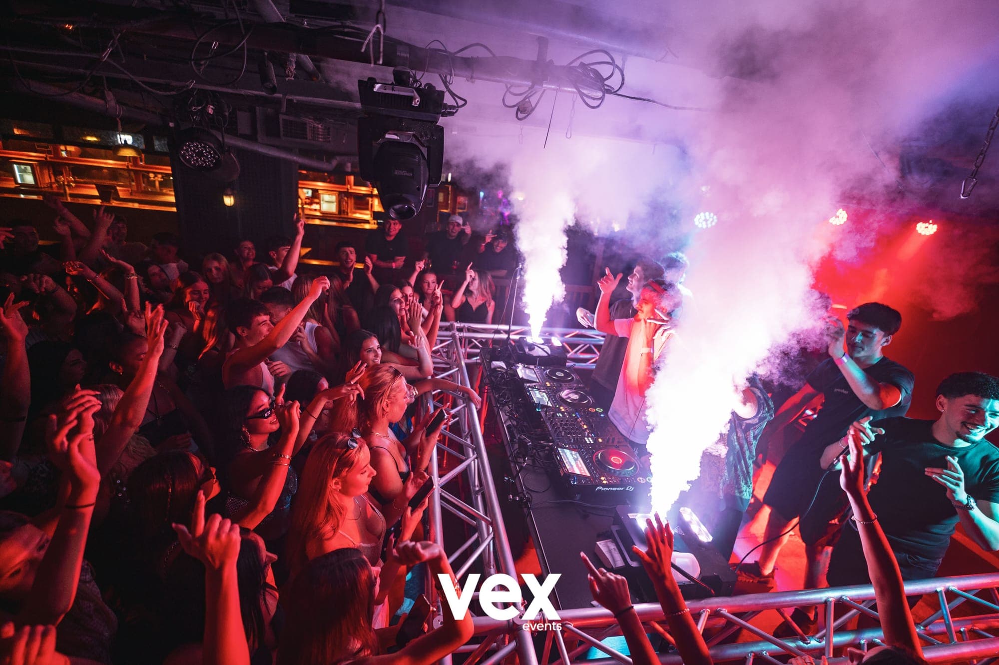 A wide indoor DJ booth surrounded by a crowd, red lighting and two tall plumes of illuminated smoke