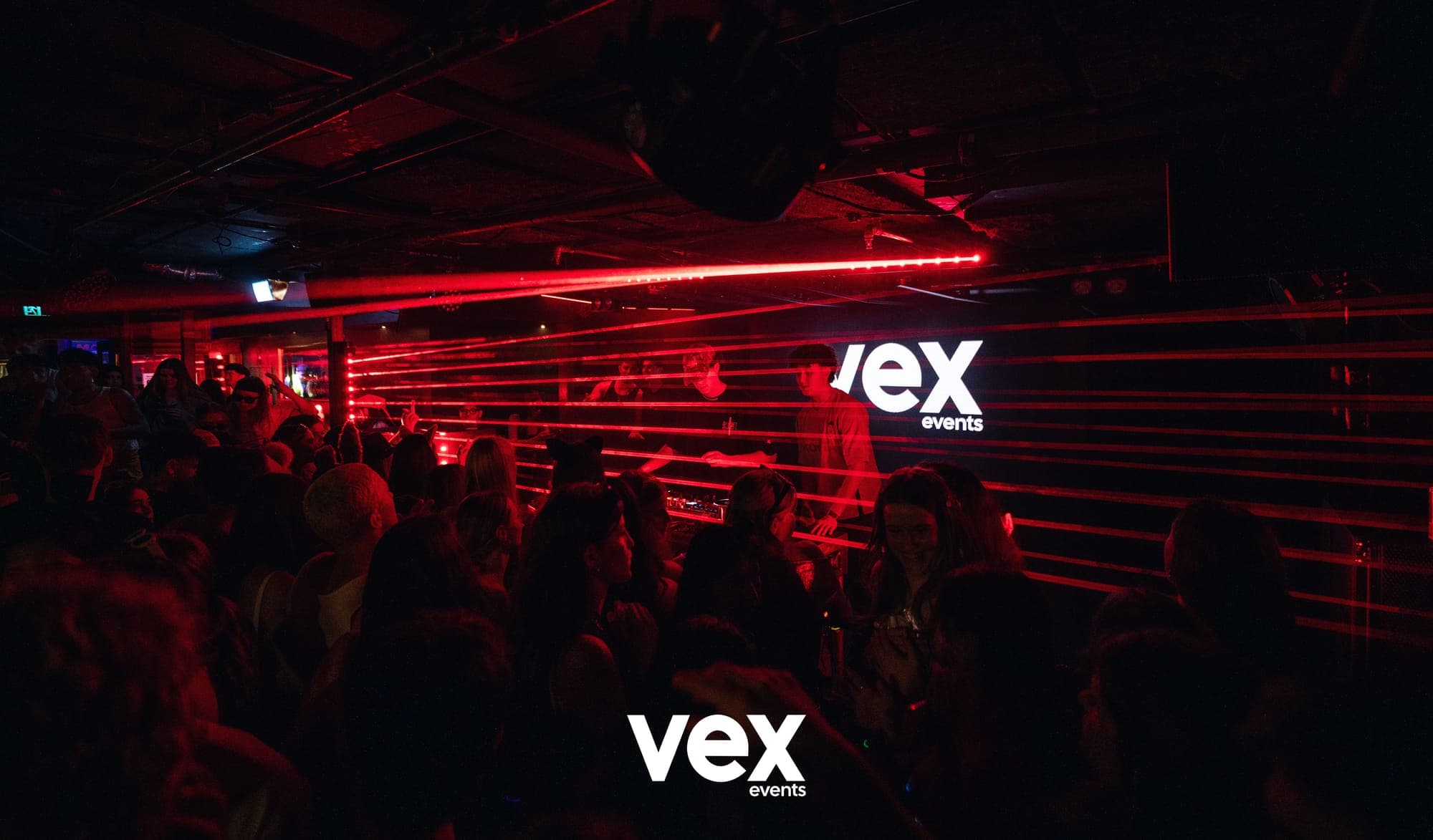 Red laser beams crossing above an indoor crowd towards a DJ booth and illuminated Vex screen