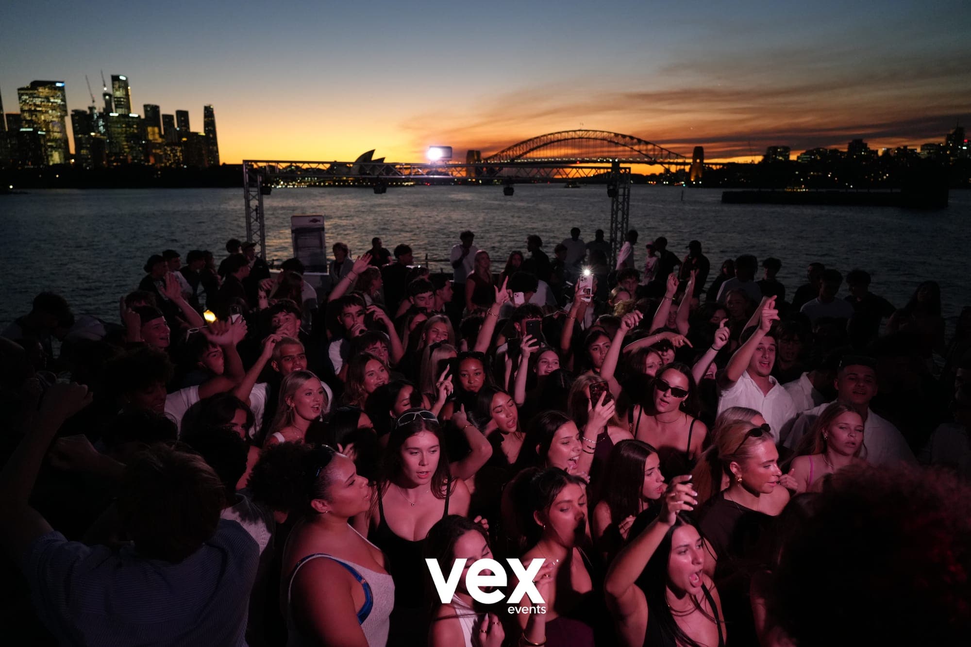 An elevated view across a boat crowd and the water towards the Sydney Harbour Bridge beneath an orange sunset